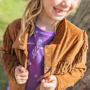 Fille souriante en gros plan portant une veste à franges brune en suède et une robe mauve à motifs western, tenant les rebords de sa veste.