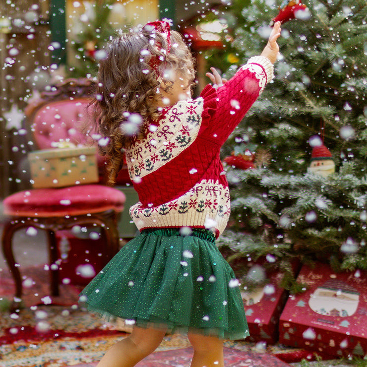 Fillette portant un chandail rouge en tricot et une jupe verte scintillante, dans une ambiance féerique de Noël près du sapin, collection Souris Mini.