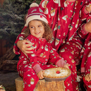 Enfant en pyjama rouge à motifs festifs assise sur une bûche, souriant dans un décor de Noël chaleureux avec sapins et tarte aux fruits
