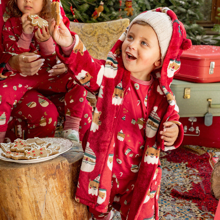 Enfant en robe de chambre rouge à motifs de Noël levant un biscuit, vêtu d’un pyjama assorti, dans une ambiance festive et chaleureuse.